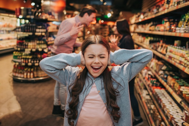 Young parents and daughter in grocery store. Girl screaming and crying. She keep ears closed. Her parents have argue and quarrel behind.