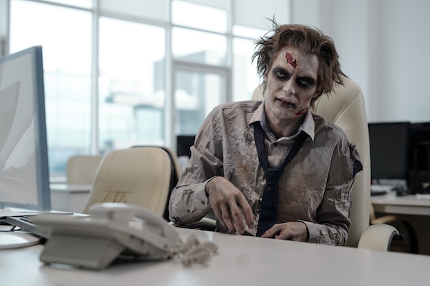 Zombie businessman sitting by desk in open space office
