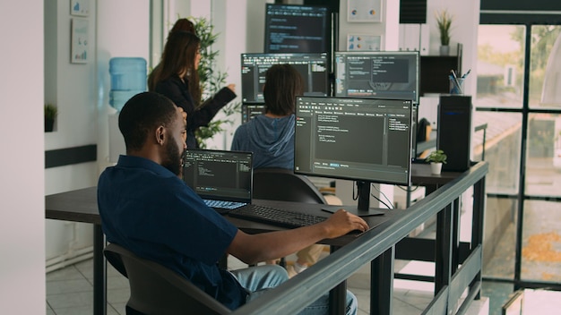 African american coder writing programming language on computer, using database code to develop server interface. System engineer analyzing html script and cloud computing in it development agency.