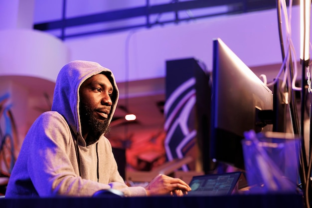 African american criminal hacking online server and stealing information Hacker in hood planning phishing attack and programming malicious software in abandoned warehouse
