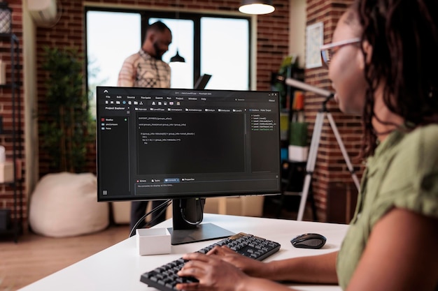 African american software developer debugging source code algorithm looking at computer screen while roommate is using laptop. Freelance coder writing software working remote from home.