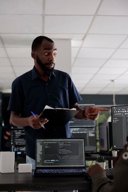 Afroamerikansk webudvikler, der analyserer HTML-script på terminalvindue og bruger serverdatabase på computer. Kodere diskuterer om programmeringssprog og cloud computing brugergrænseflade.