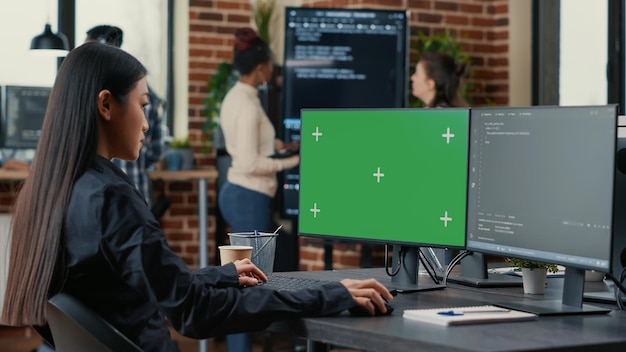 Asian programer writing code in front of computer with green screen chroma key mockup in software development agency. Coder computing big data with team colleagues working online cloud computing.