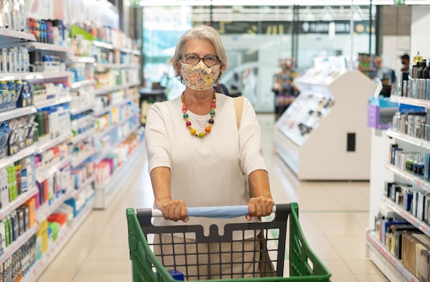 attractive senior woman makes purchases in the supermarket by pushing the cart wearing a protective face mask