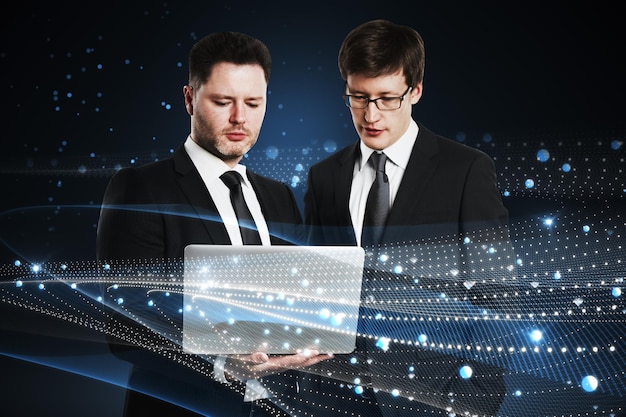 Attractive young european businessmen with laptop standing on abstract glowing blue metaverse background Digital world teamwork and technology concept