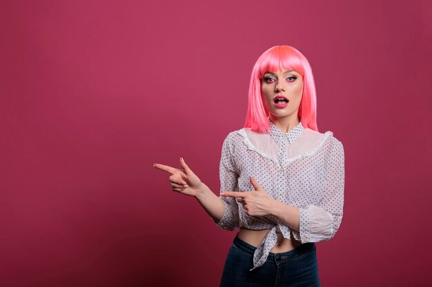 attractive young woman with pink hair pointing sideways in studio, indicating left or right directions in front of camera. recommending that way with raised arms and index fingers.