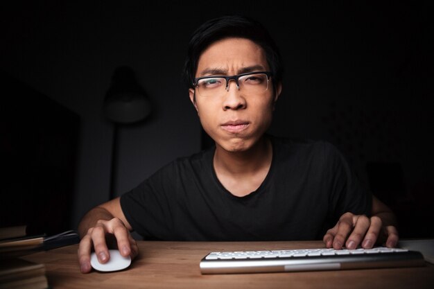 Concentrated asian young man in glasses working with computer in dark room