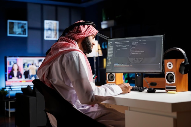 Dedicated software programmer wearing traditional Arabic clothing is seated at his home office desk examining database code on computer. Muslim man checking the programming language on the pc monitor.
