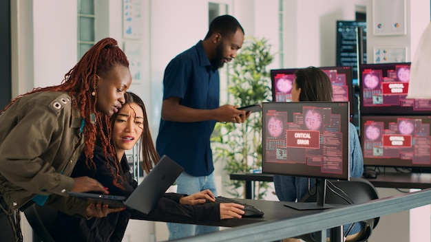 Diverse group of engineers having computer hacked and system crash on multiple monitors, reading critical error message flashing on screen. Security breach alert and hacking warning. Handheld shot.