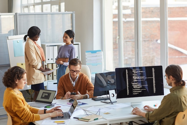Diverse team of IT developers using computers and programming code while collaborating on project in software production studio