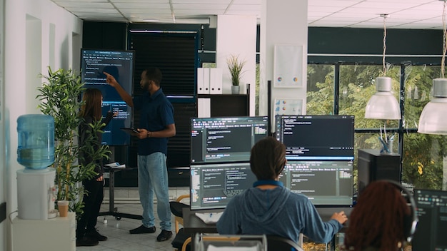 Diverse team of software engineers analyzing code on wall screen tv, using digital tablet and working with artficial intelligence. Coders parsing cloud computing project with big data.