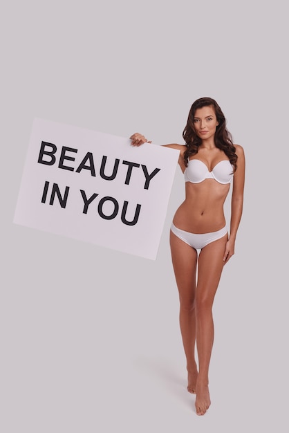 Every woman should know this.  Full length of attractive young woman holding a poster and looking at camera with smile while standing against grey background