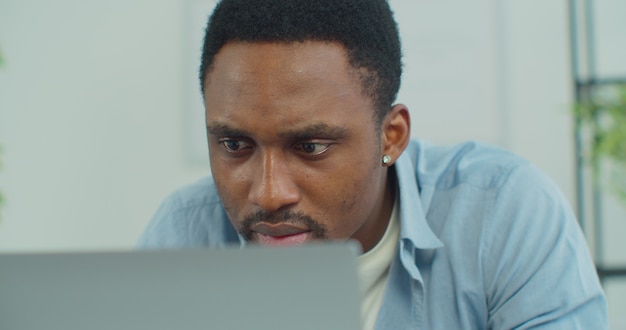 Face of handsome african man surfing internet chatting use laptop at home freelancer man working remotely looking at screen of computer pc