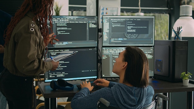 Female developer analyzing program code and talking to asian engineer with chronic disability, collaborating on new interface development. Wheelchair user and app developer programming language.