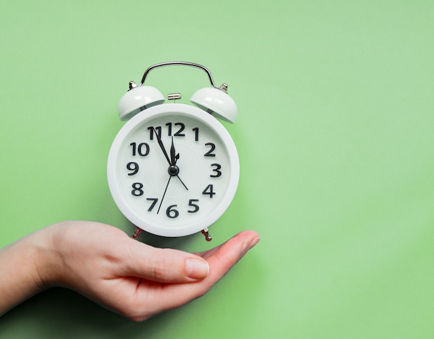 Female hand holding an alarm clock on pastel green background.