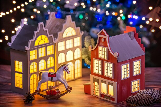 festive composition of a lamp in the form of an old european house, a toy horse, fir branches and cones against the background of defocused garlands