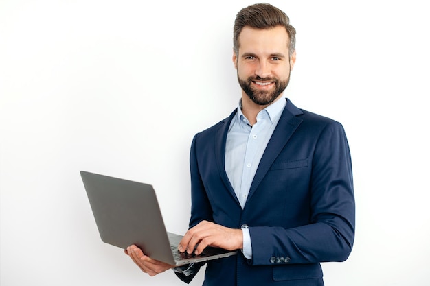Friendly attractive caucasian bearded business man in a suit programmer IT specialist holding opening laptop in hands standing on isolated white background looks at camera smiling