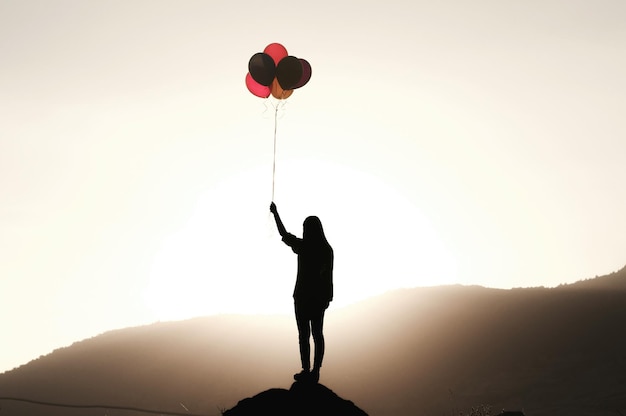 Full length of silhouette woman holding colorful balloons on cliff against mountain