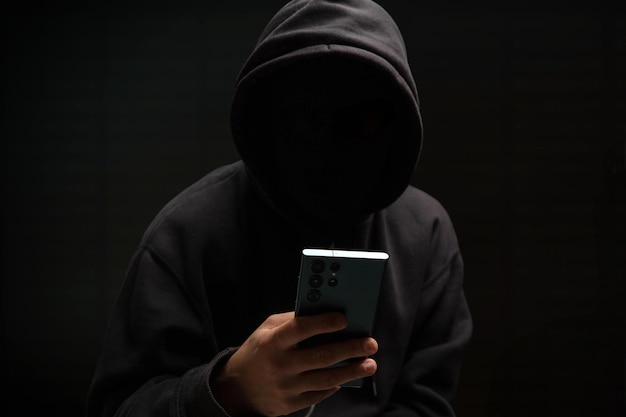 Hacker in black hood holding a cellphone Scam cyber crime concept