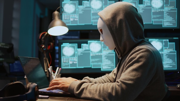impostor with mask hacking computer server at night, working on laptop and multiple monitors to steal it information or data. masked hacker with hidden identity, security breach. handheld shot.