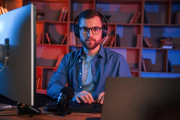 In glasses Man is sitting by a computer indoors at home Neon lighting