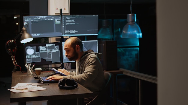 IT-webbutvecklare jämför datafiler på urklippspapper och flera skärmar för att programmera nytt servergränssnitt. Utvecklar html-skript på terminalfönstret, kodar databas. Handhållen bild.