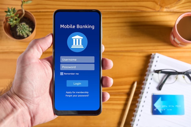 Male hand holding mobile phone with online banking app interface on the screen login page with username and password boxes with credit card and accessories on wooden table blurred in the background