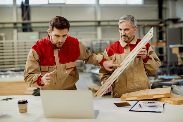Male workers using computer while comparing measurements of a piece of wood at carpentry workshop