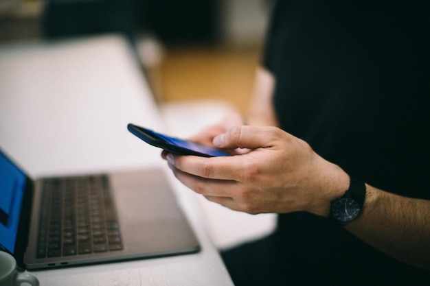 Man i mellansegment som använder mobiltelefon vid bärbar dator på bord på kontoret