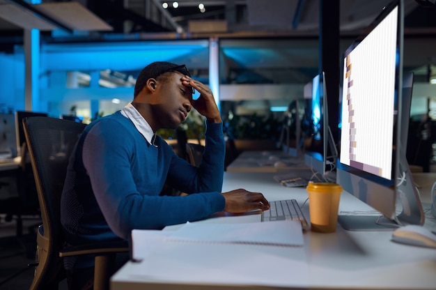 Manager sover ved bordet, natkontorlivsstil. Mandlig person ved bærbar computer, mørkt interiør, moderne arbejdsplads