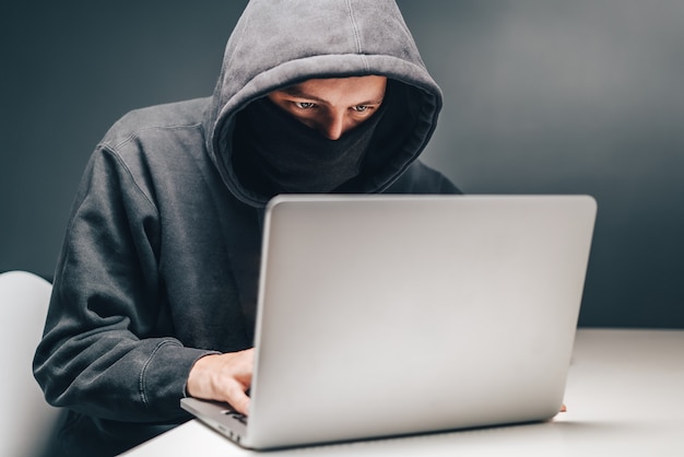 Mandlig hacker med hætte og skjult ansigt får adgang til personlige oplysninger på bærbar computer i mørket
