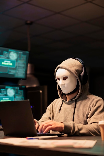 Masked scammer hacking computer network at night, working on laptop and multiple monitors to steal IT information. Hacker with hidden identity acting dangerous, server security breach.