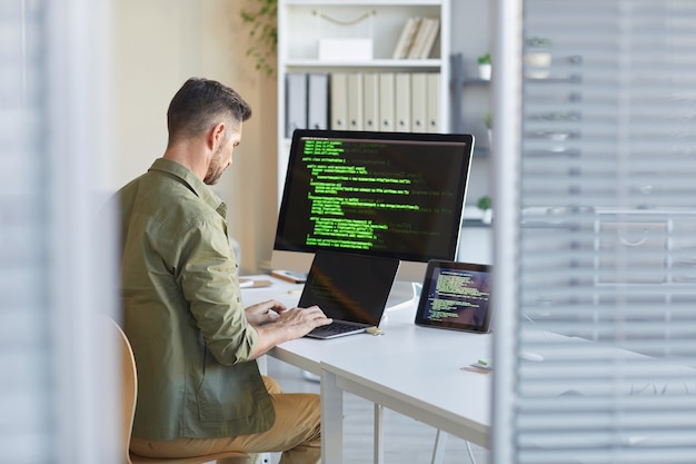 Mature technician sitting at his workplace in front of computer monitor and typing on laptop at IT office
