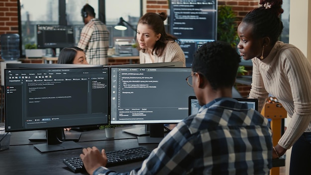 Mixed team of artificial intelligence cloud programers talking about programming in front of multiple screens compiling code. Programmers doing teamwork looking at machine learning algorithms.