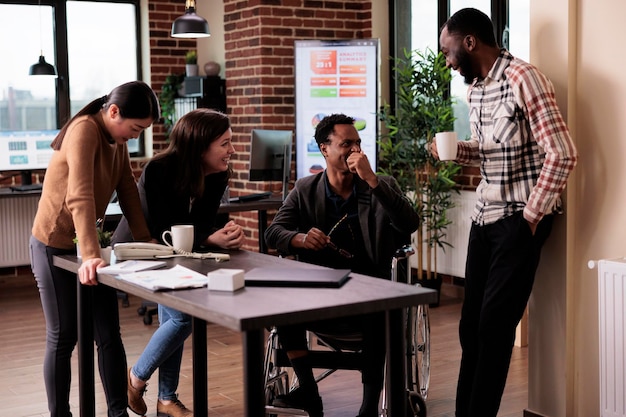 Multiethnic team of happy people and paralyzed man taking break from work in business office. Male employee wheelchair user suffering from physical health condition laughing with colleagues.