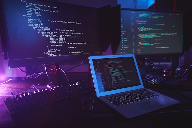 olika datorutrustning med programmeringskod på skärmarna på bordet i ett mörkt rum, cybersäkerhetskoncept, kopieringsutrymme