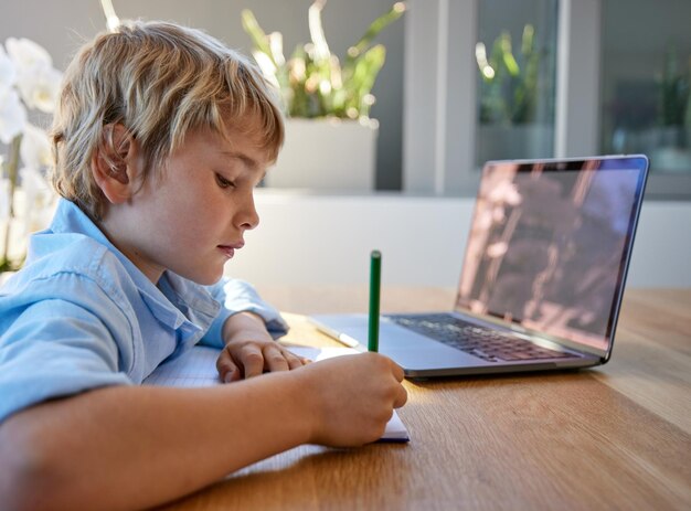 Outdoor laptop and boy writing with homework for education elearning or school work at home Online studying and young child at table with notebook for development growth or knowledge in backyard