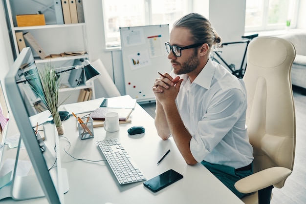 Overvejer næste skridt. Topvisning af eftertænksom ung mand i skjorte, der arbejder med computer, mens han sidder på kontoret
