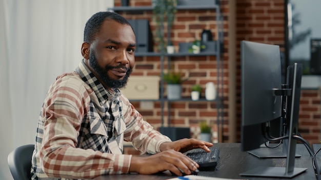 Portrait of programer typing programming code on computer keyboard turning head and smiling while colleague walks by in background. Software engineer sitting at desk writing code feeling confident.