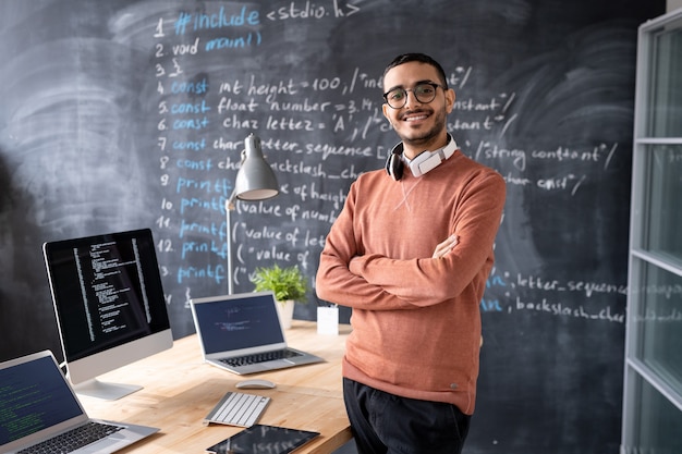 Portrait of satisfied young middle-eastern computer programmer with headphones around neck standing with crossed arms in coding studio