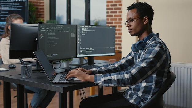 Programer thinking while touching beard and fixing glasses while typing on laptop sitting at desk with multiple screens parsing code. Focused database admin working with team coding in the background.