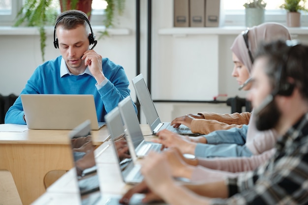 Serious young contact center employee adjusting microphone in headset while working with customers complaints