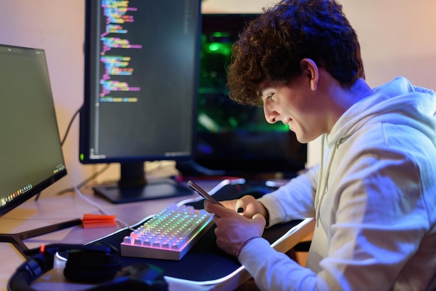 Side view of smiling young male gamer looking at screen of mobile phone while sitting in illuminated armchair at table with gaming PC