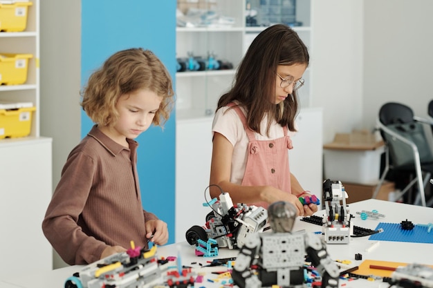Side view of two pupils standing by table with equipment for robot constructing