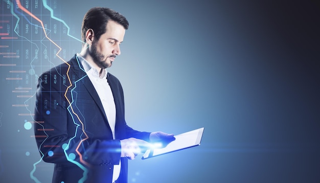 Side view of young european businessman using pad with growing blue forex chart hologram on blurry gray background with mock up place Banking invest and financial growth concept