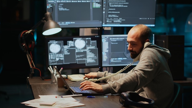 Software developer talking on landline phone at night, answering phone call in office. Male programmer using telephone with cord to chat and work on online cloud computing, artificial intelligence.