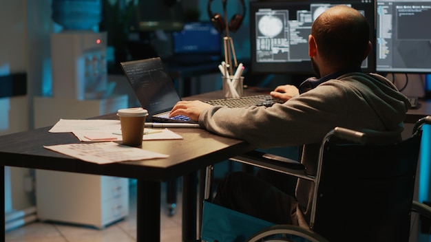 Software developer with impairment working on app programming, using laptop and multiple screens. Male IT coder with chronic disability in wheelchair developing new user interface.