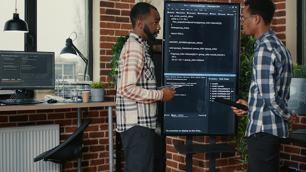 Team of software engineers leaving after analyzing source code on wall screen tv comparing errors using digital tablet. Coders passing coworker programmer writing code for artificial intelligence.