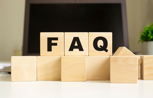 the word faq is written on wooden cubes lying on the office table in front of a laptop.