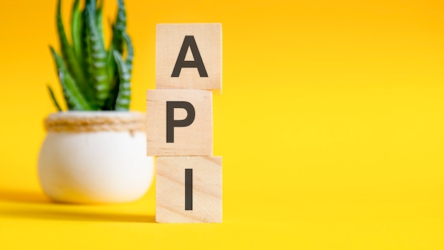 Tre trækuber med bogstaver - API, på gult bord, plads til tekst til højre. Frontvisningskoncepter, blomst i baggrunden. API- forkortelse for APPLICATION PROGRAMMING INTERFACE
